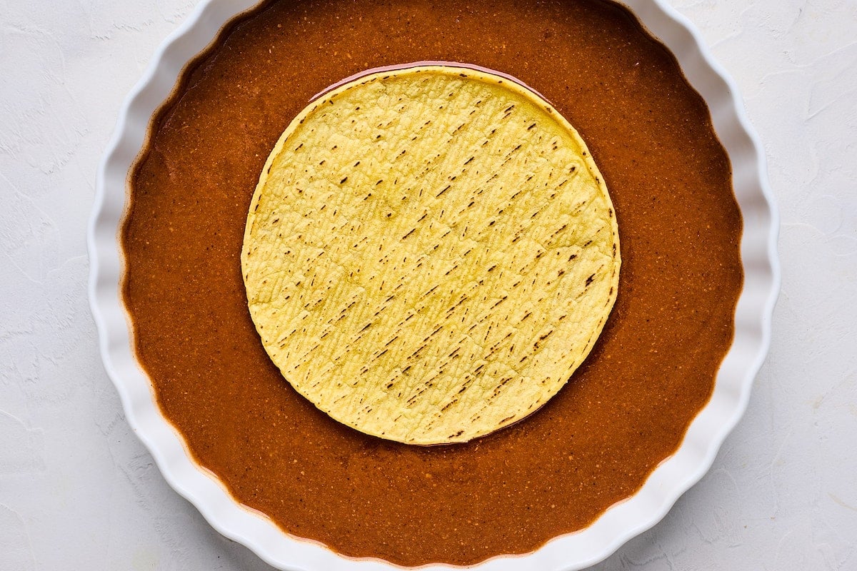 corn tortilla being dipped in enchilada sauce in shallow bowl.