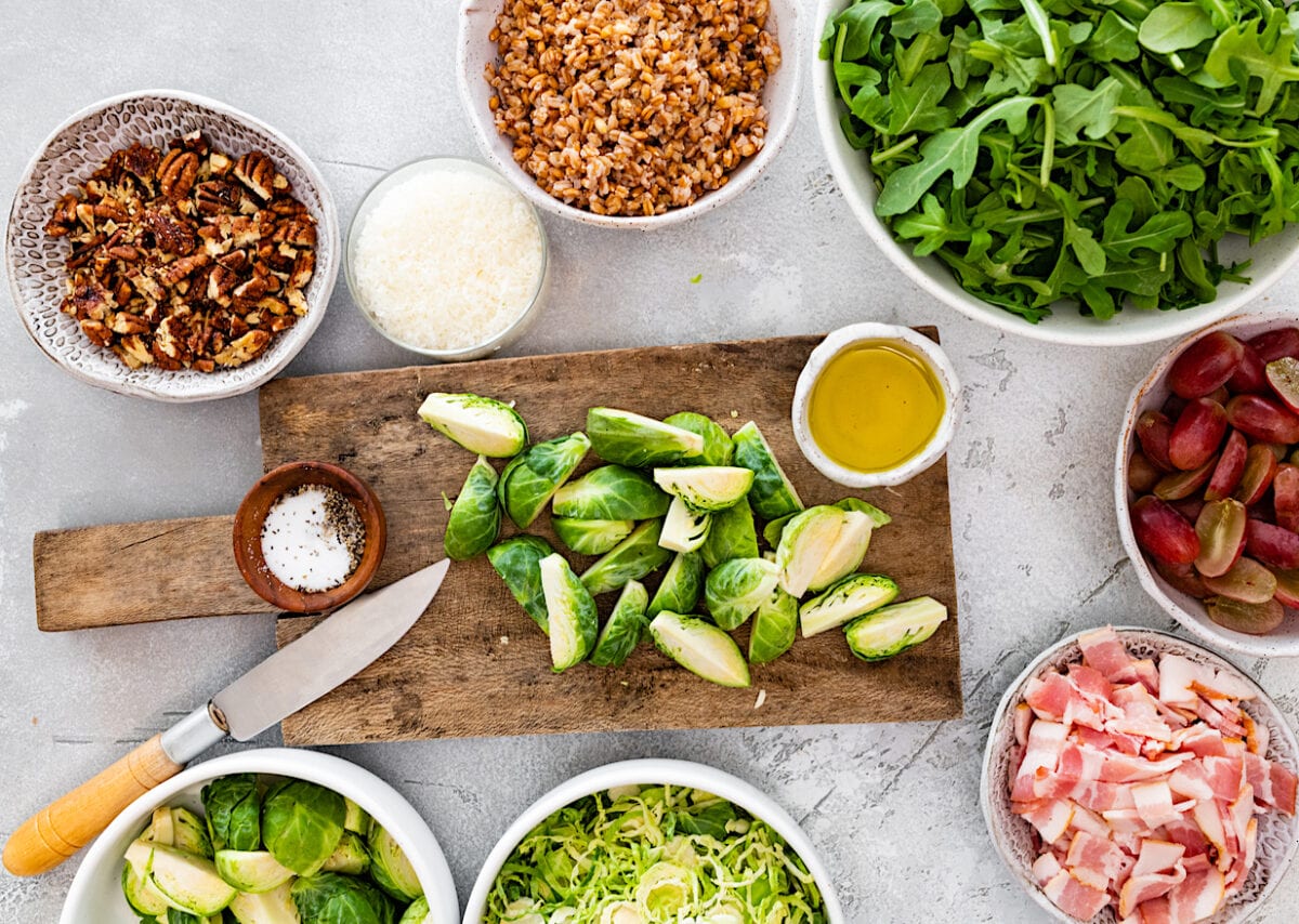 brussels sprouts being cut on a wood cutting board with salad ingredients in bowls.