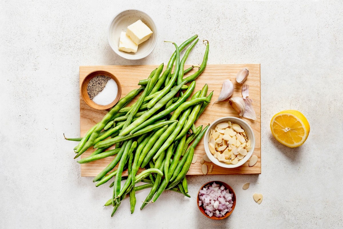 green beans on wood cutting board with salt, pepper, almonds, shallot, lemon, and butter to make green beans almondine.