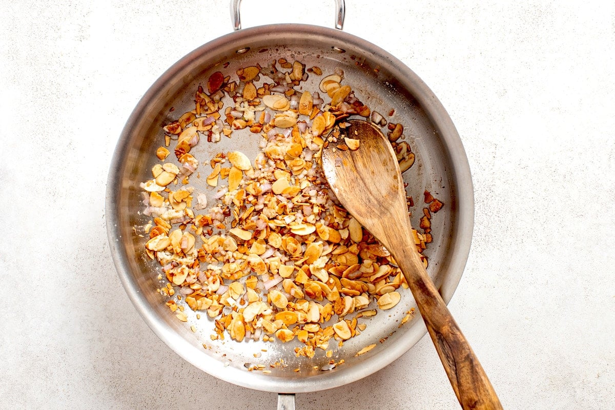 sliced almonds being cooked in skillet with wooden spoon.
