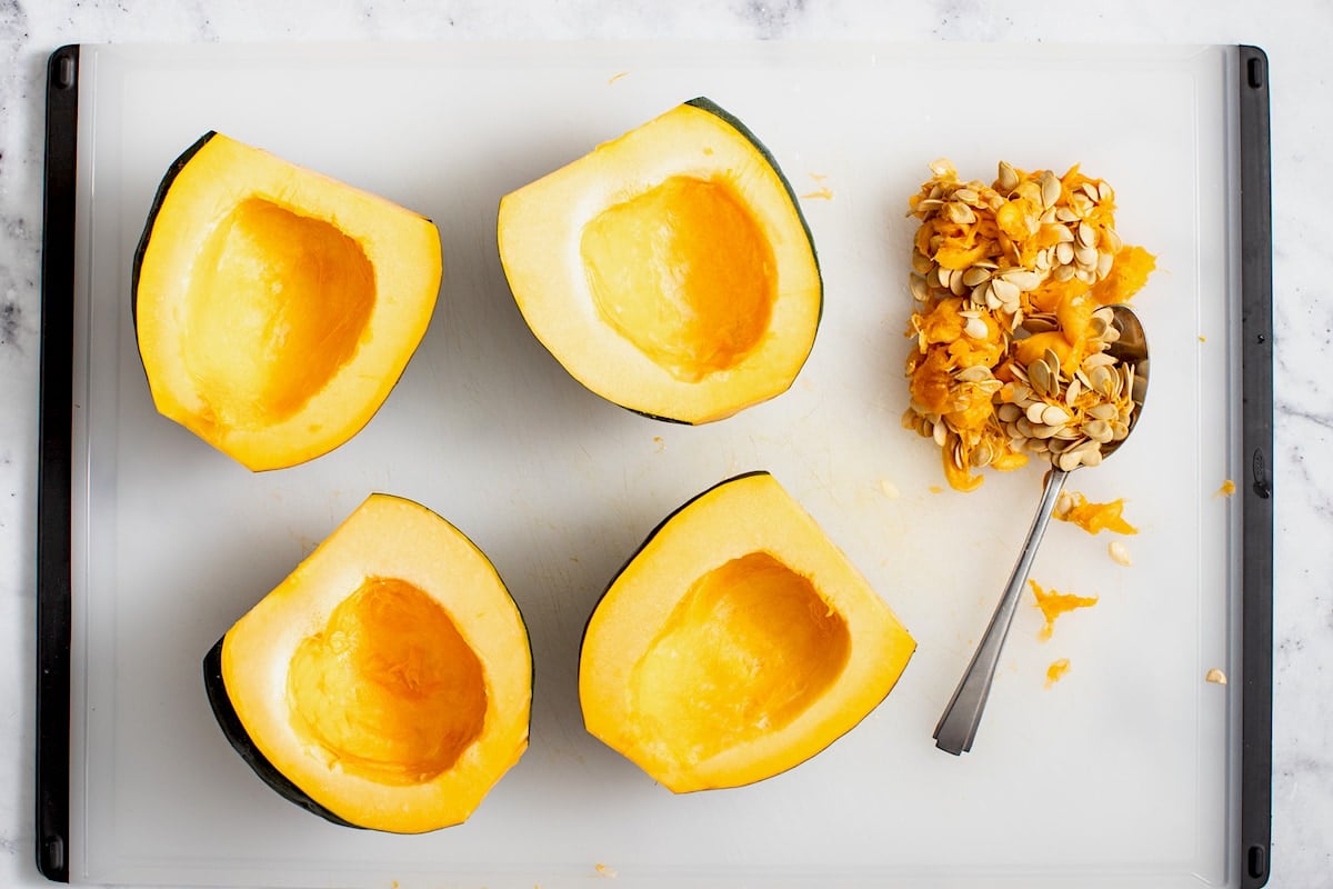 seeds being removed from acorn squash halves with spoon on cutting board.