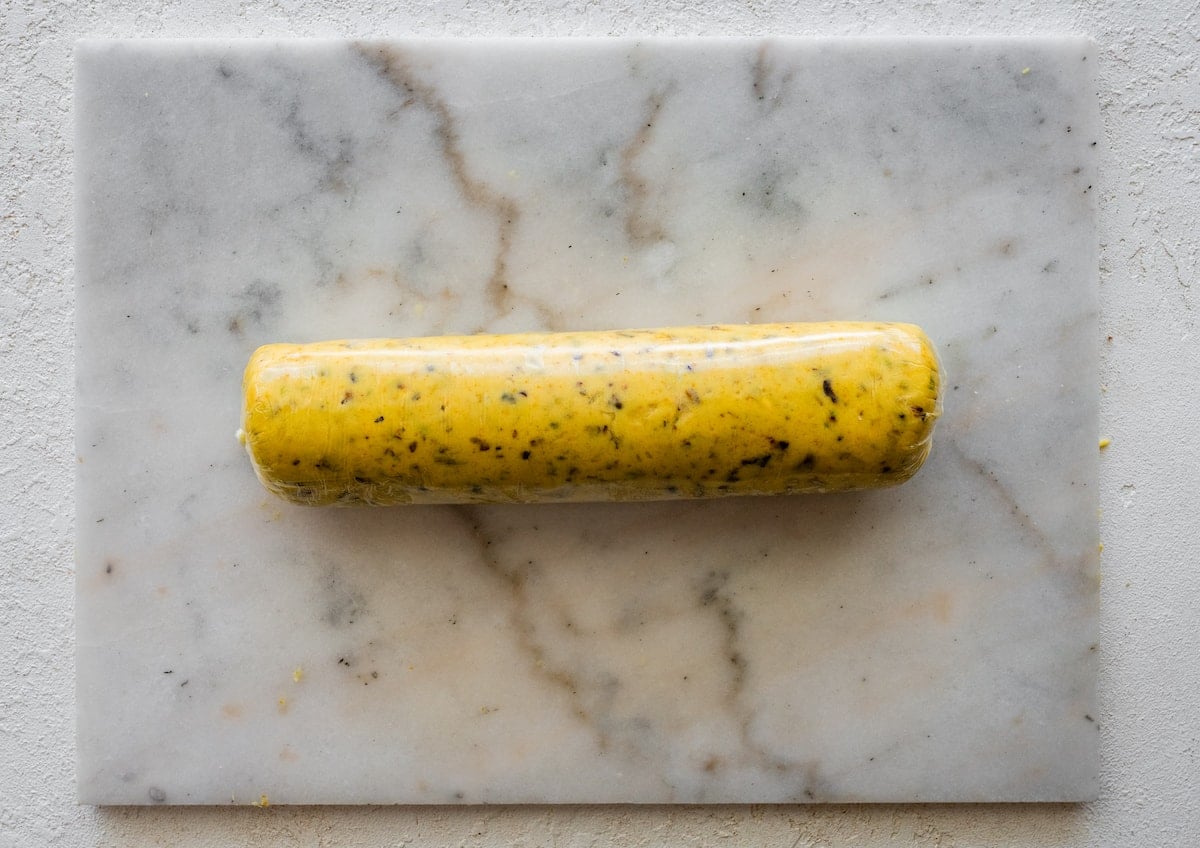 cranberry orange pistachio dough rolled in log in plastic wrap on marble surface.