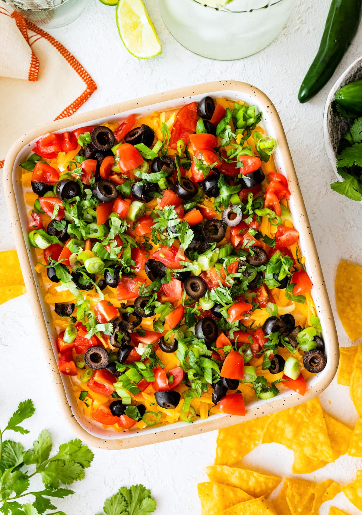 7 Layer Dip in pan with cheese, tomatoes, olives, green onion, cilantro and chips on the side.