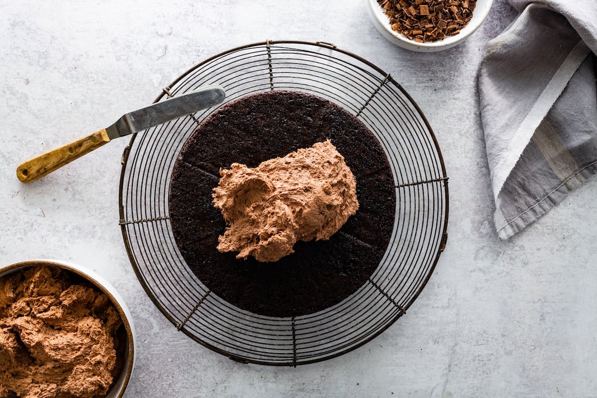 chocolate cake being frosted with chocolate frosting on cooling rack.