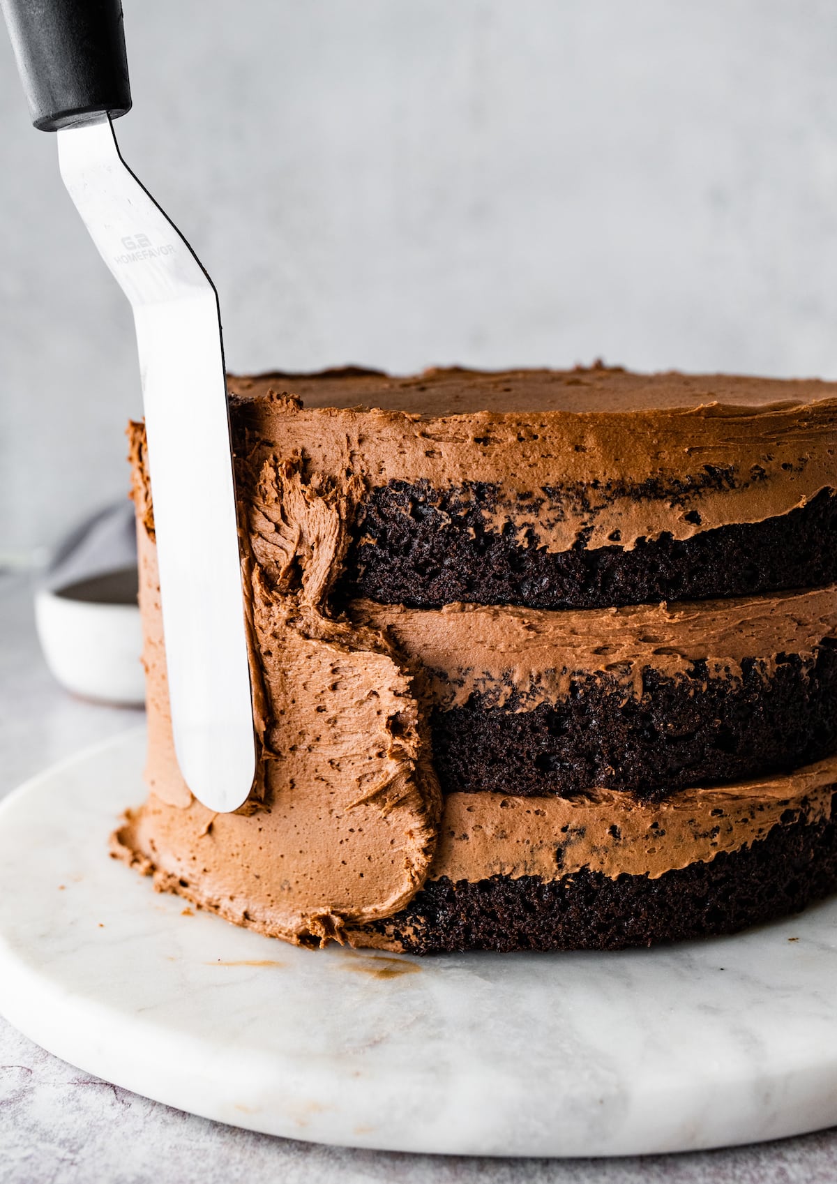 layered chocolate cake being frosted with an offset spatula.