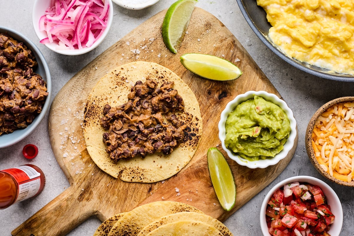 assembling breakfast tacos with beans, scrambled eggs, guacamole, pico de gallo, cheese, and pickled red onions.