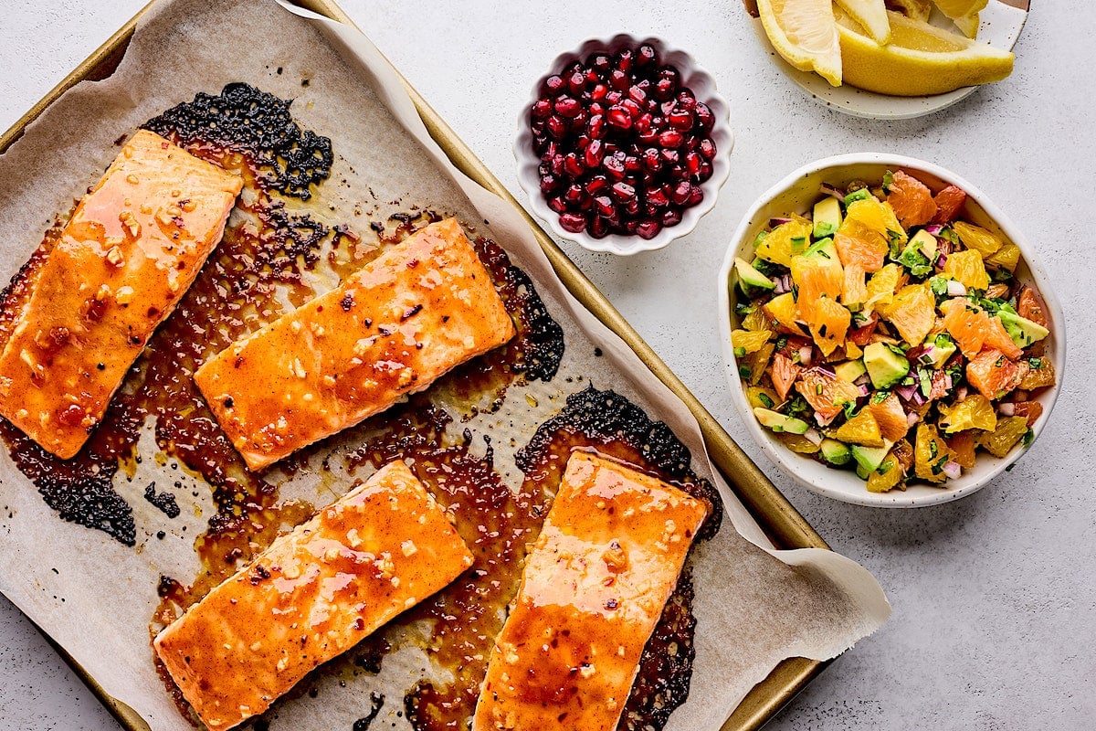 honey mustard salmon on baking sheet with citrus salsa and pomegranate arils on bowls.