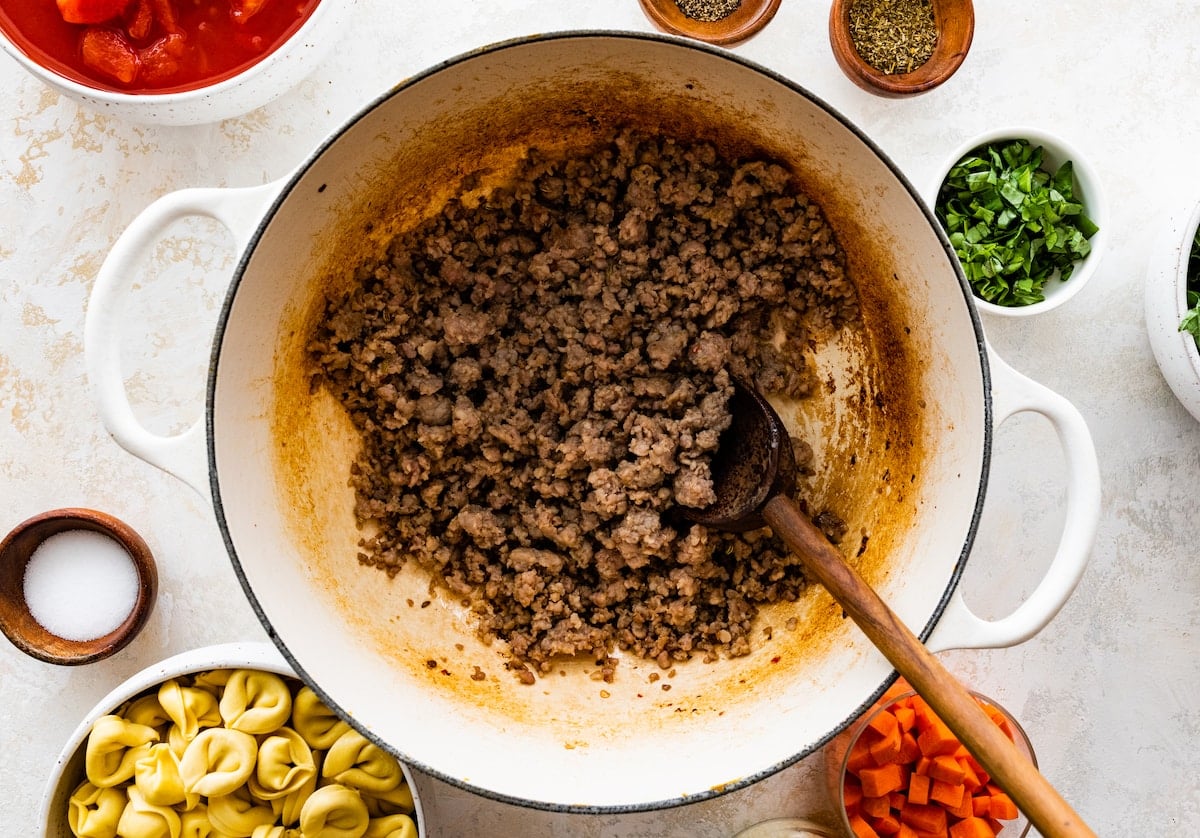 Italian sausage cooking in large white pot with wooden spoon.