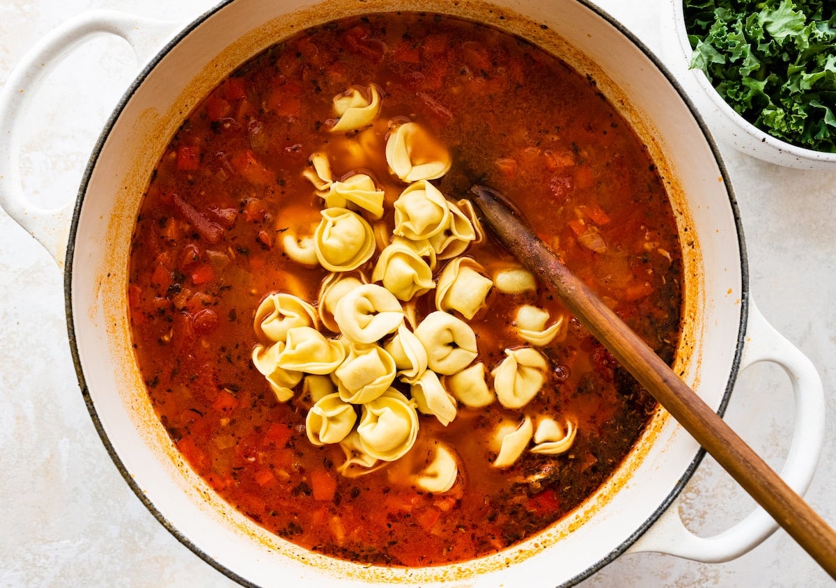 Italian sausage tortellini soup cooking in a pot with wooden spoon.