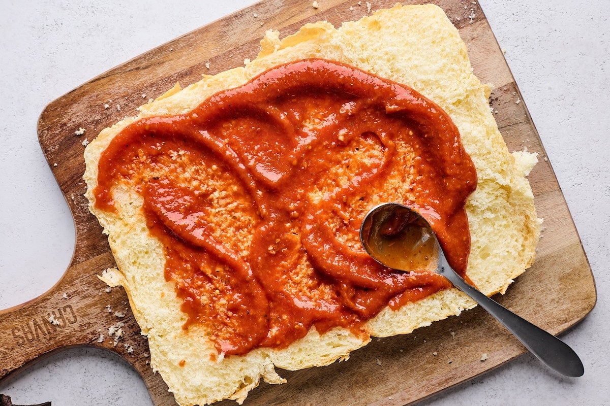 pizza sauce being spread on top layer of Hawaiian sweet rolls with spoon.