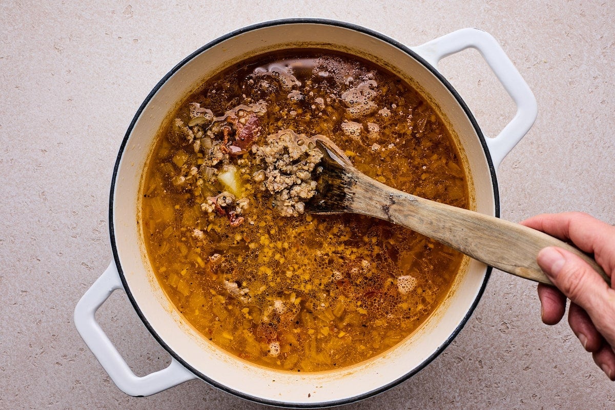 stirring sausage and bacon into zuppa toscana soup in large white pot.