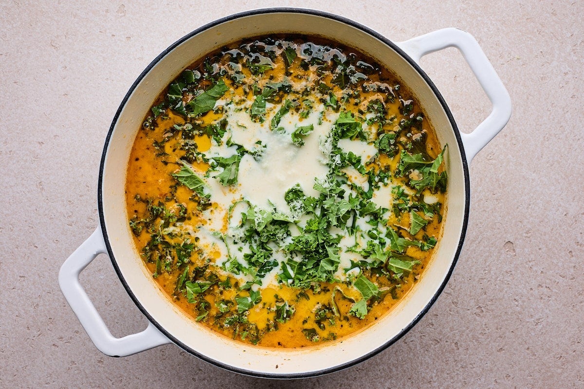 heavy cream and kale being added to zuppa toscana soup in large white pot.