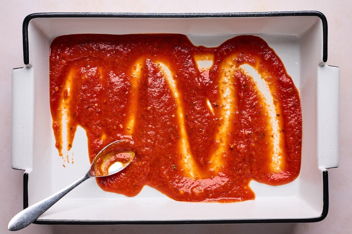 spreading marinara sauce in baking dish with spoon.