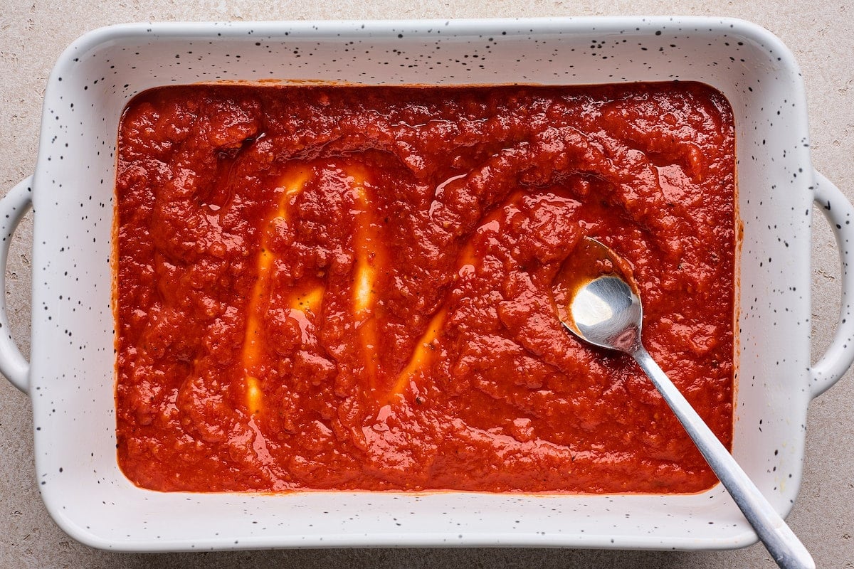 marinara sauce spread at the bottom of a baking dish with spoon.