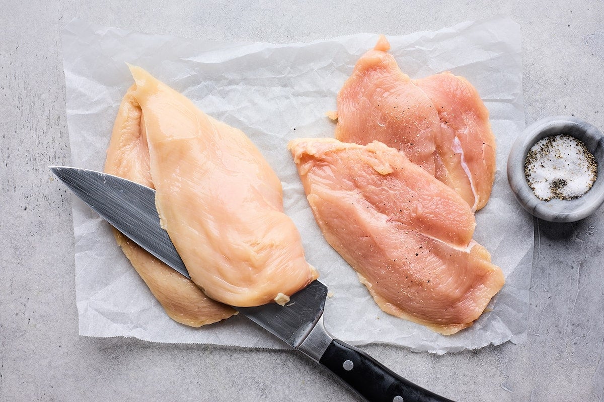 boneless skinless chicken breasts being cut in half with sharp knife to make spinach artichoke chicken.