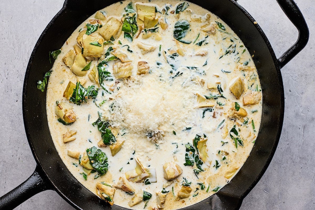 Parmesan cheese being added to creamy spinach artichoke sauce.