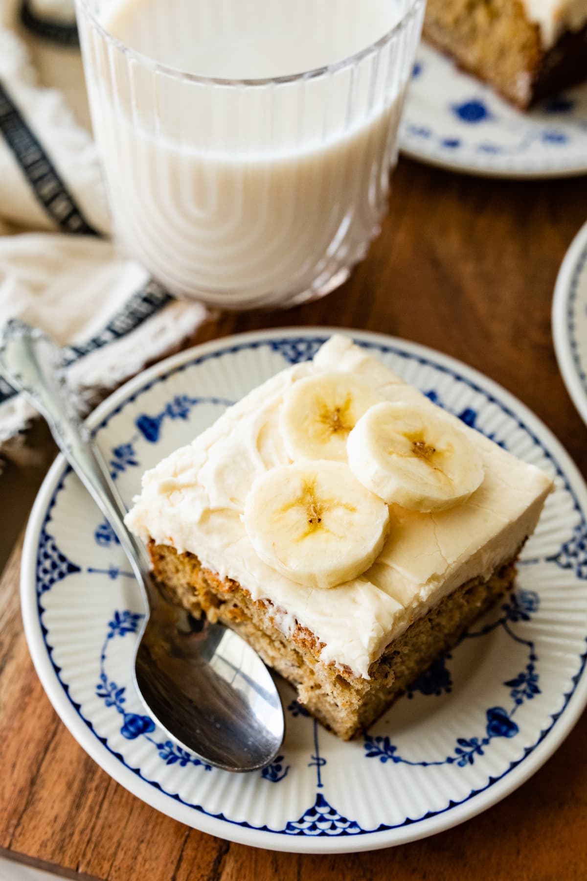 piece of banana cake with cream cheese frosting on plate with spoon and glass of milk.