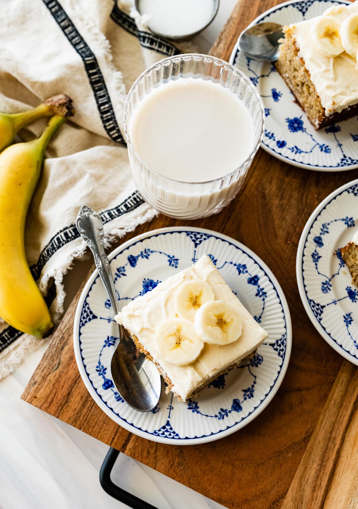 pieces of banana cake with cream cheese frosting and fresh banana slices on plates with glass of milk.