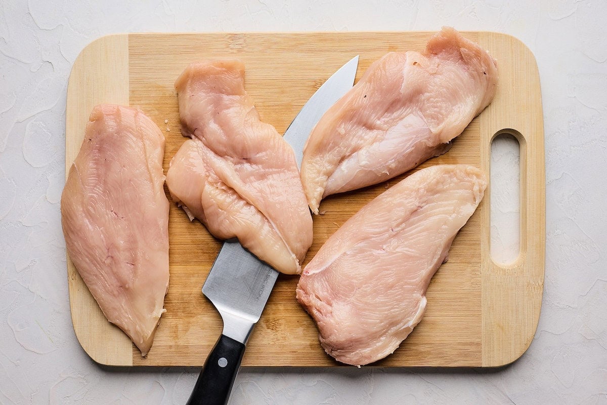 chicken breasts being cut in half with a sharp knife on a cutting board.