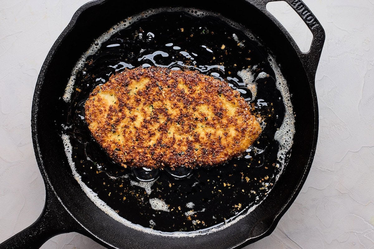 chicken Milanese frying in oil in cast iron skillet.