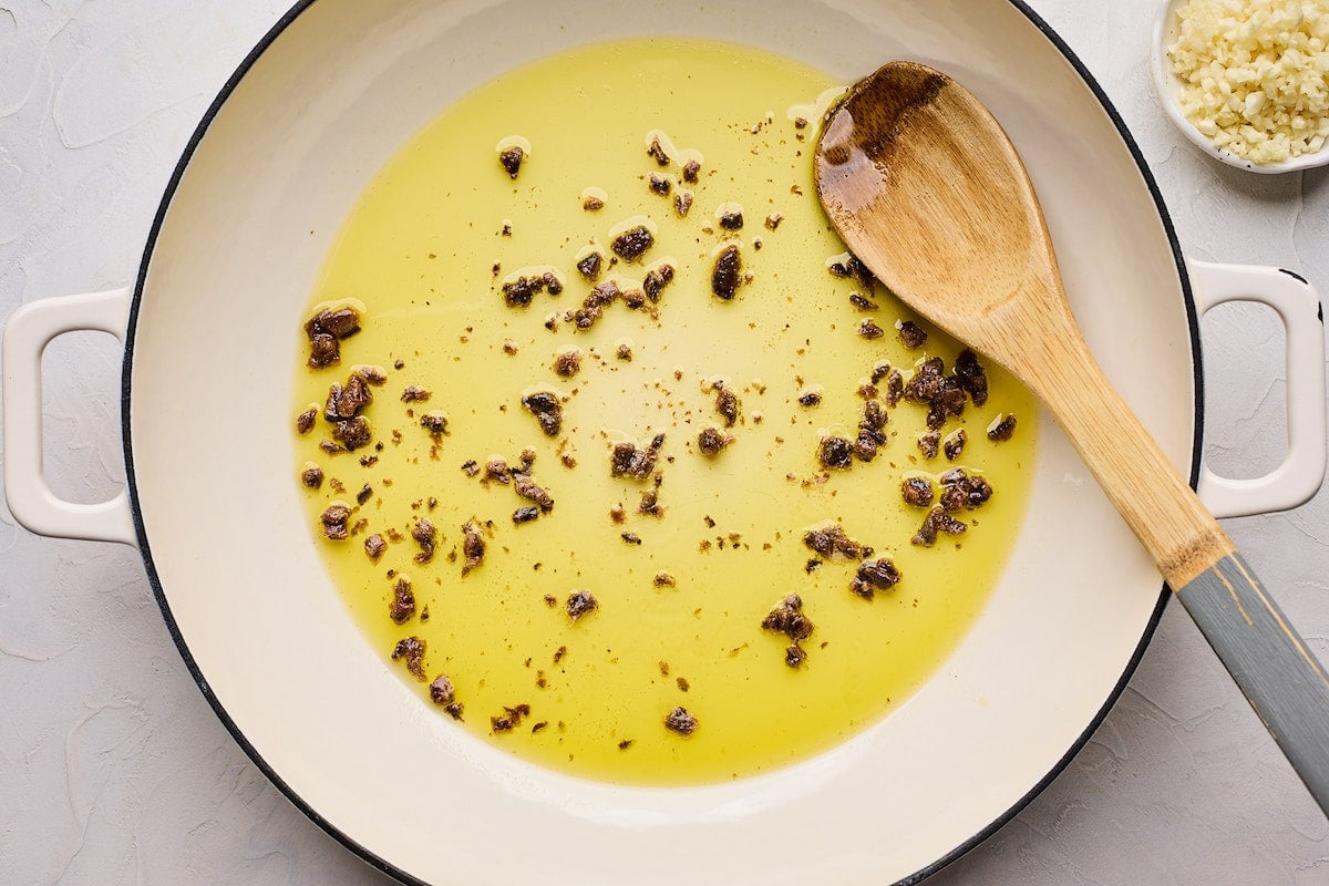 olive oil with anchovies in skillet with wooden spoon to make pasta puttanesca.