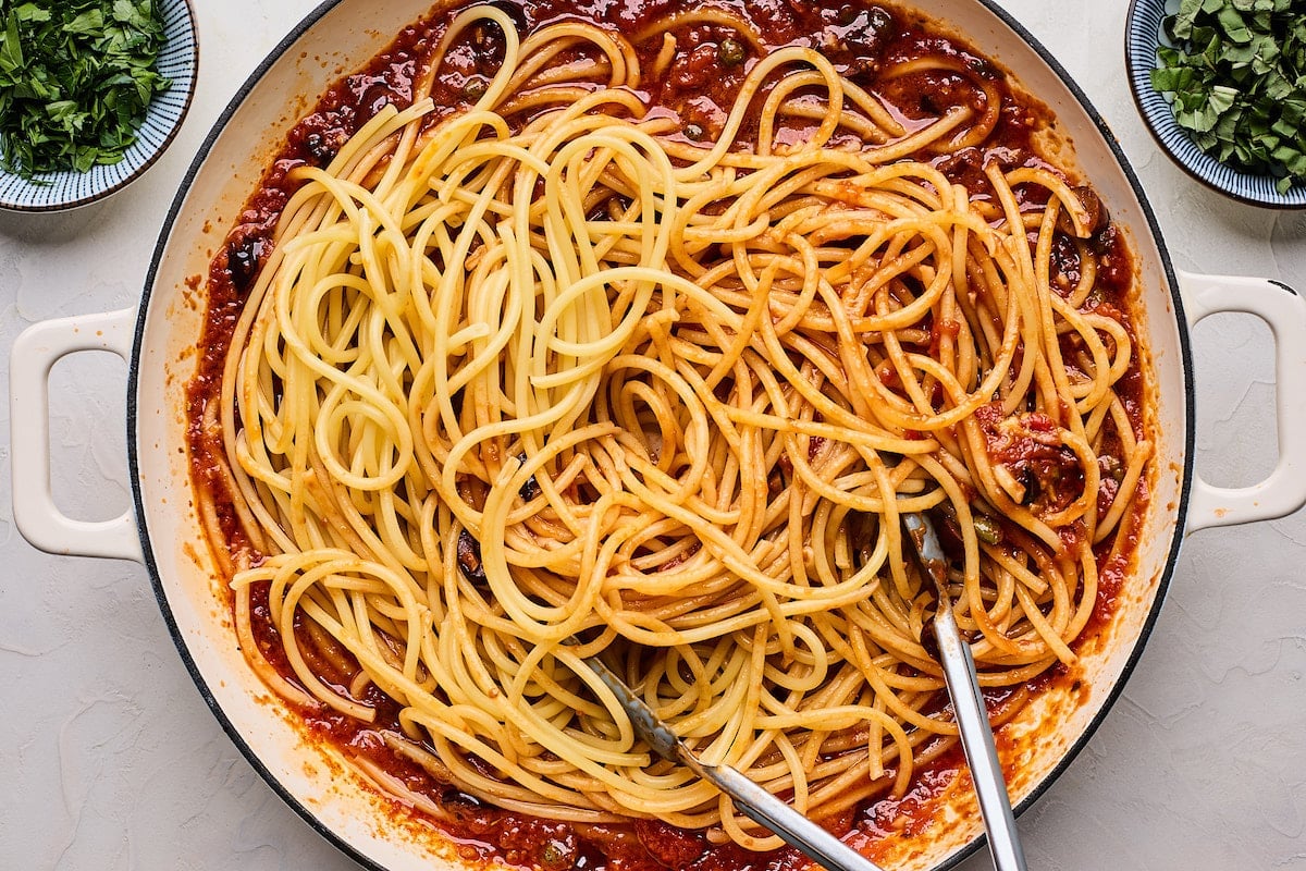 spaghetti in skillet with tongs and puttanesca sauce.