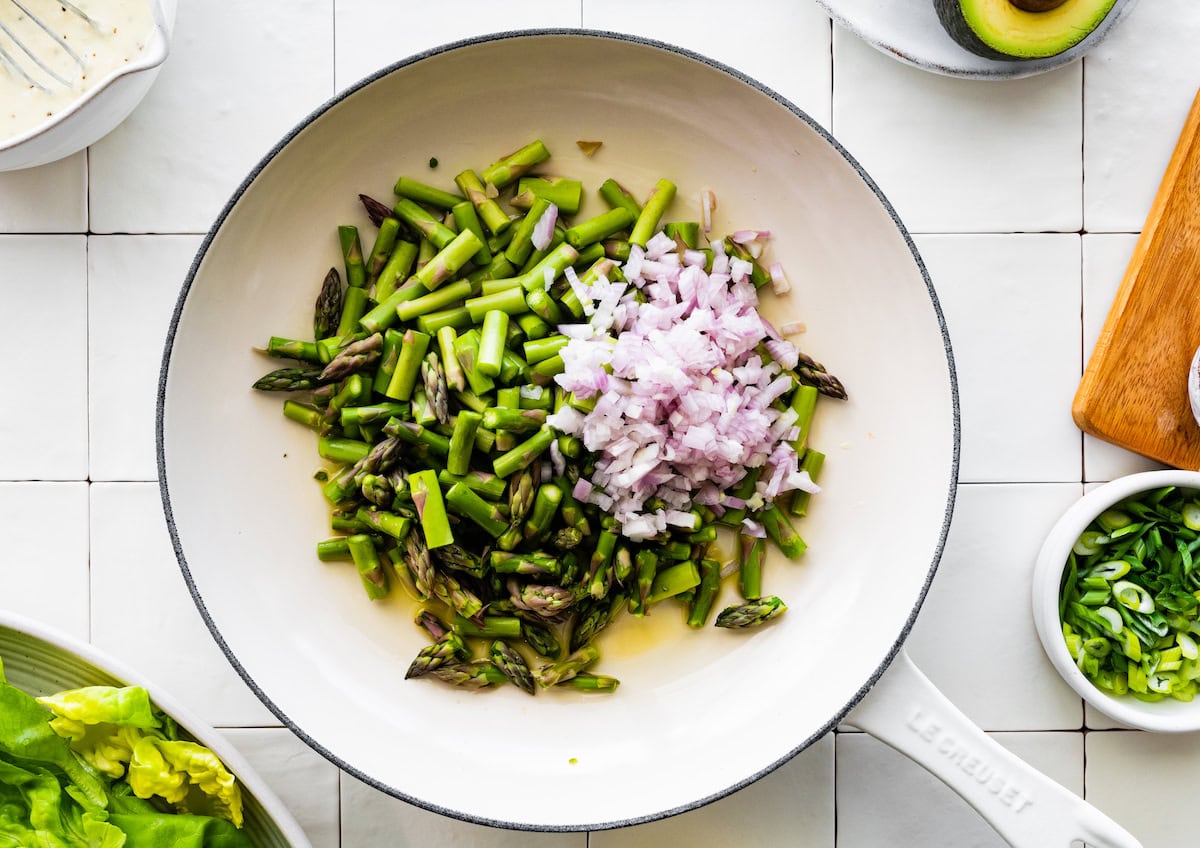 shallot and asparagus cooking in oil in white skillet.