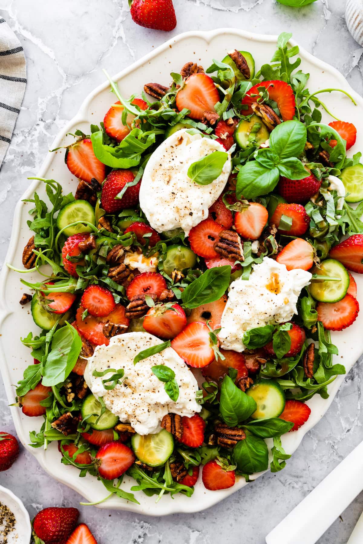 strawberry burrata salad on platter with strawberries, cucumbers, candied pecans, burrata, and fresh basil.