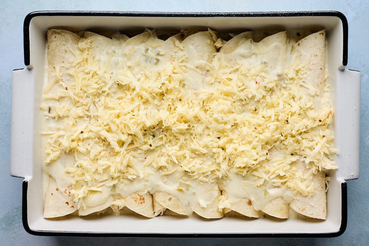 rolled up white chicken enchiladas in pan covered with sauce and shredded cheese.