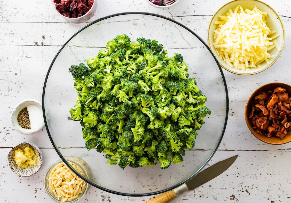 chopped broccoli in mixing bowl with other ingredients in bowls to make broccoli salad.