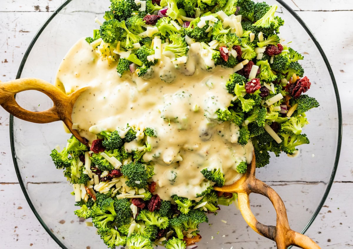 creamy broccoli salad dressing being tossed over broccoli salad in mixing bowl with salad servers.