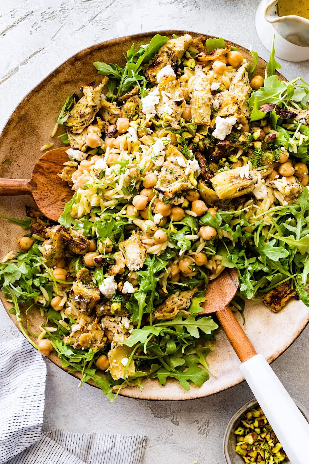 roasted artichoke chickpea orzo salad with arugula being tossed in bowl with wood salad servers.