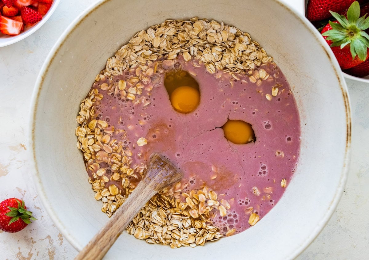 oats, eggs, and strawberry milk in mixing bowl with wooden spoon to make strawberry baked oatmeal.