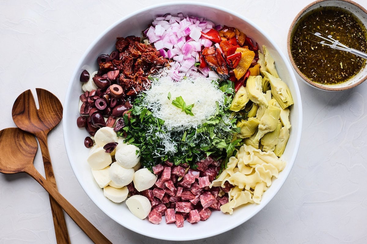 salami, pasta, artichoke hearts, peppers, onion, sun dried tomatoes, olives, mozzarella cheese, parmesan cheese, and herbs in serving bowl to make Italian pasta salad.