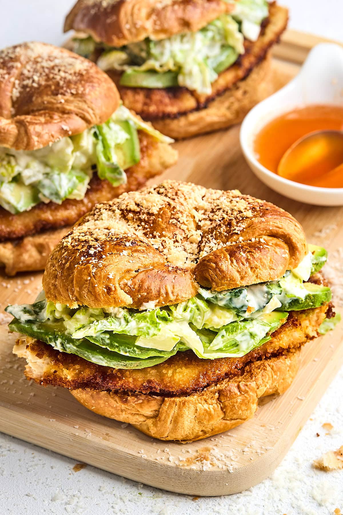 crispy chicken croissant sandwich with lettuce, avocado, and parmesan cheese on wood cutting board.