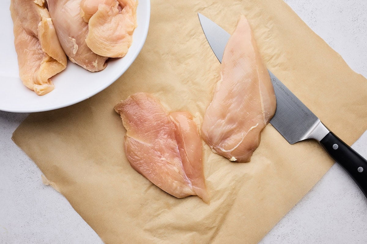 A sharp knife cutting chicken breasts in half widthwise.