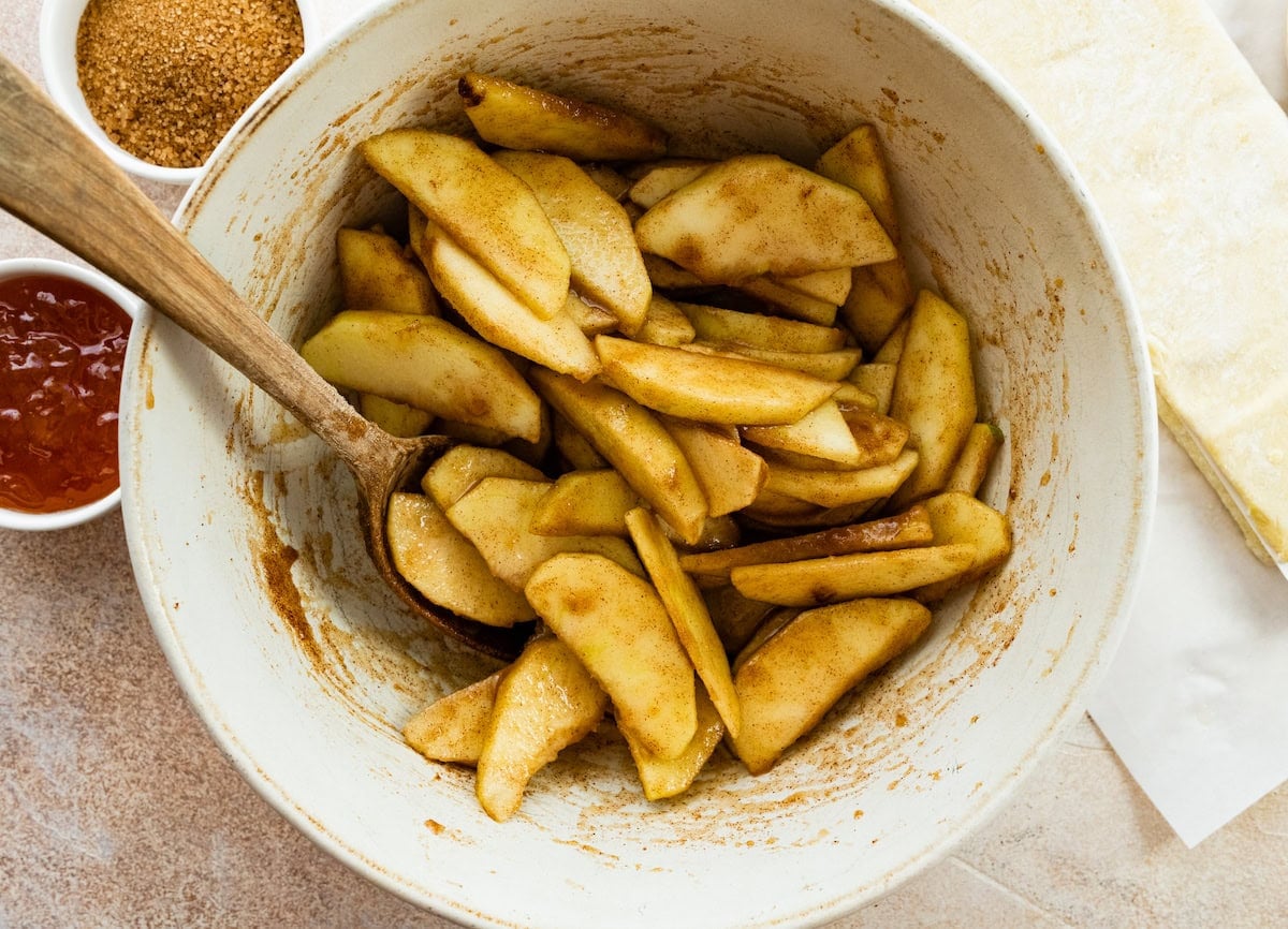 apple slices tossed in sugar and cinnamon in mixing bowl with wooden spoon.