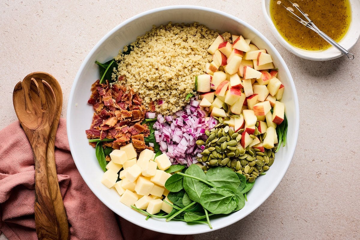 spinach, quinoa, apple, pepitas, white cheddar cubes, bacon, and red onion in serving bowl.