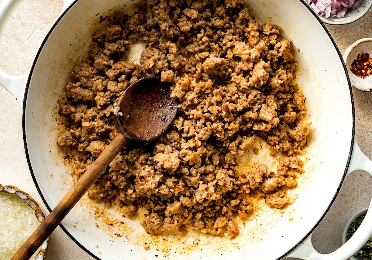 Italian sausage cooking in skillet with wooden spoon.
