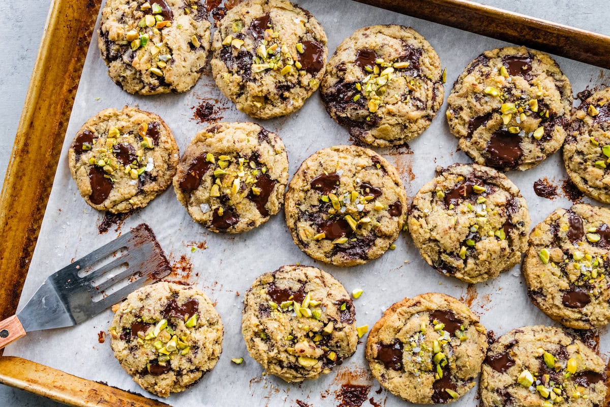 dark chocolate chunk pistachio cookies on baking sheet with parchment paper and spatula.
