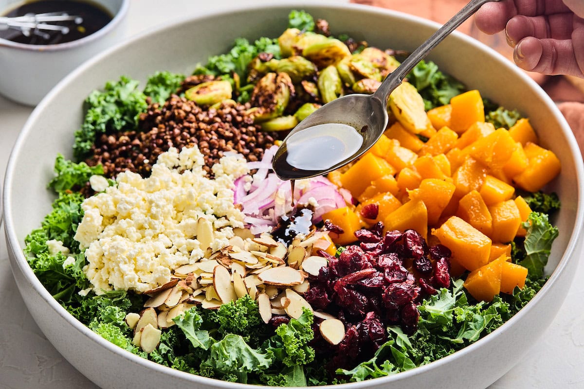 maple mustard dressing being spooned over kale salad with roasted vegetables and crispy lentils.