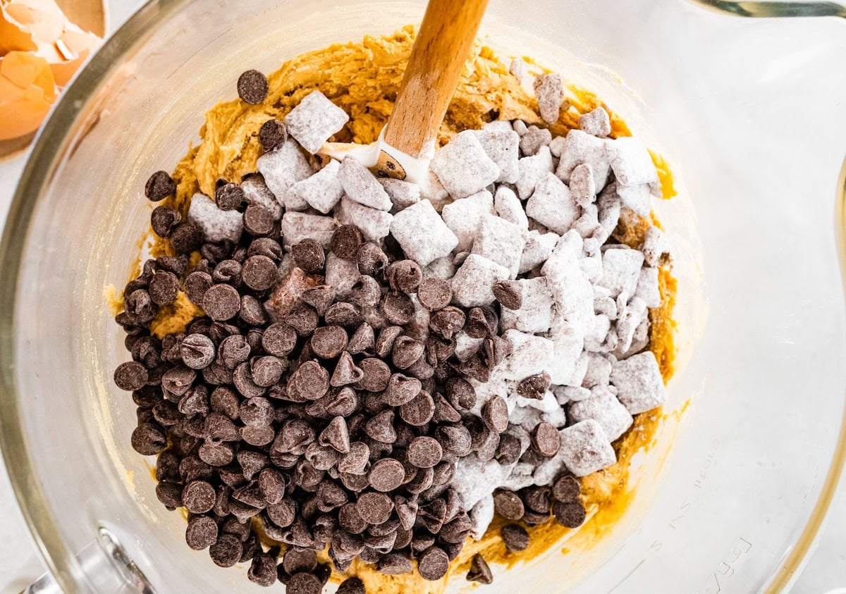 puppy chow and chocolate chips being stirred into cookie dough with spatula in mixing bowl.