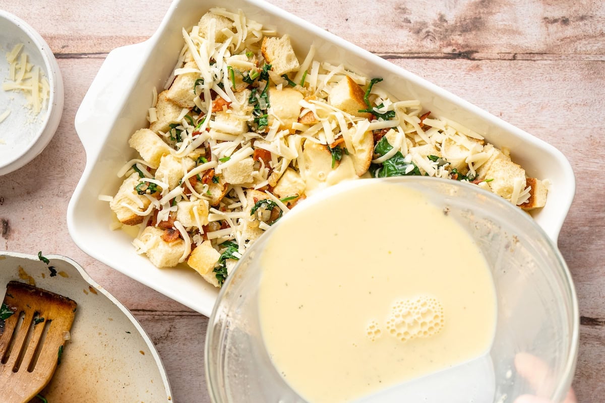 egg mixture being poured over bread cubes, bacon, spinach, and cheese to make breakfast strata.