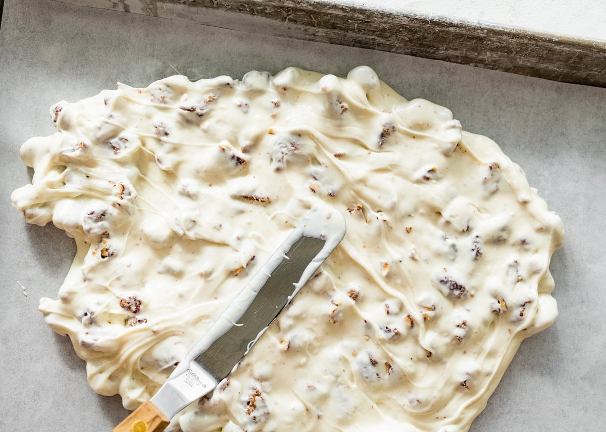 melted white chocolate and candied pecans being spread on baking sheet with parchment paper to make bark.