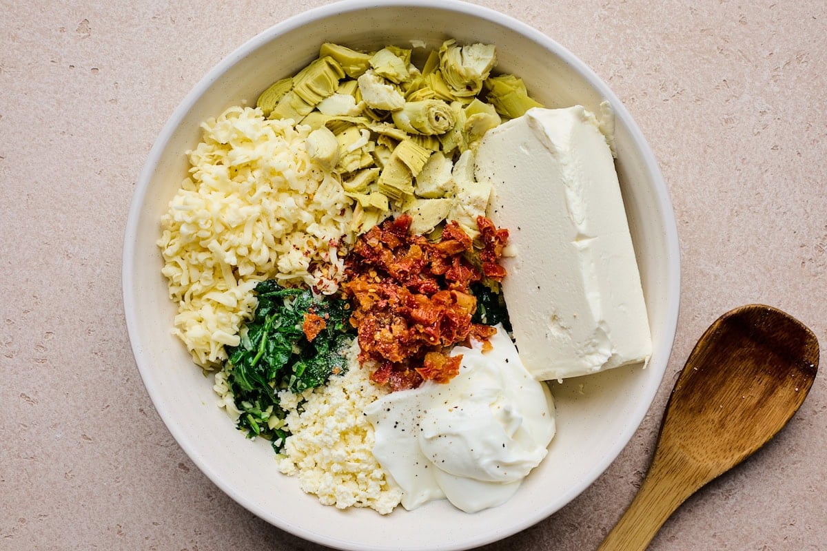 cream cheese, greek yogurt, feta cheese, mozzarella cheese, spinach, artichokes, and sun-dried tomatoes in a mixing bowl with wooden spoon.