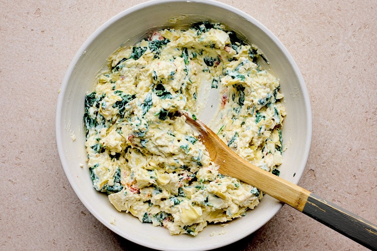 creamy spinach artichoke sun-dried tomato dip being combined with spoon in mixing bowl.
