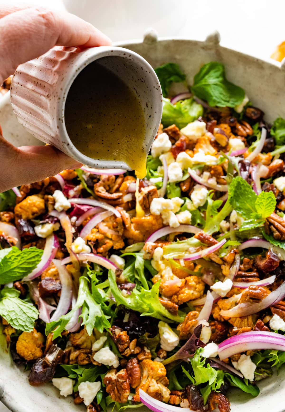 pouring orange maple vinaigrette over a roasted cauliflower salad with dates, pecans, and red onion.