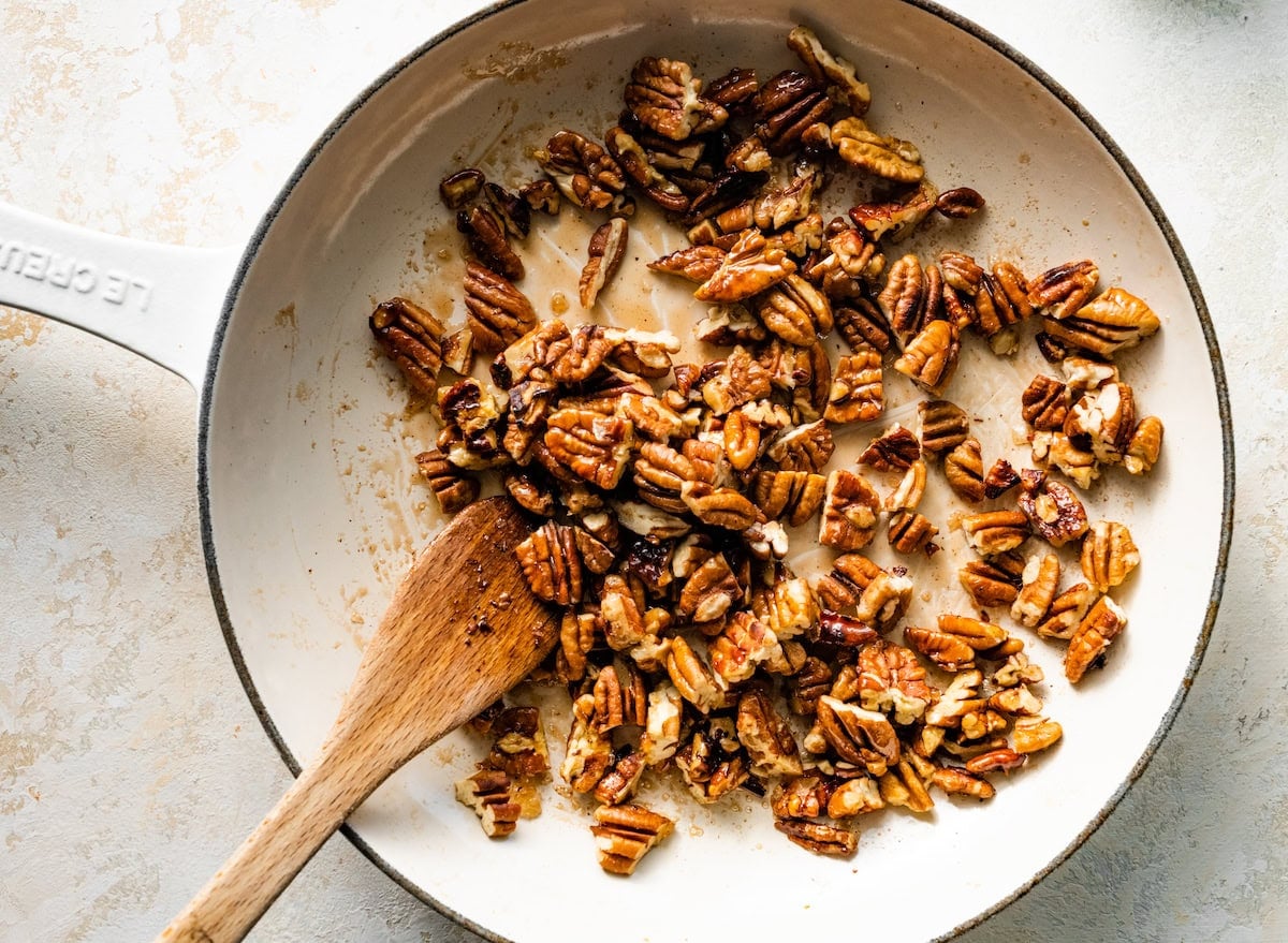 salted maple pecans cooking in skillet with wooden spoon.