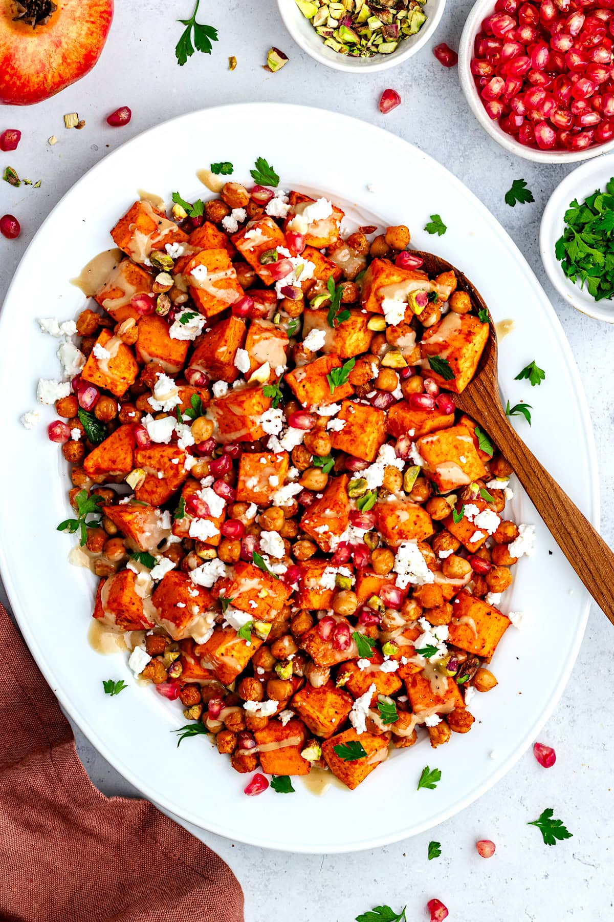 Roasted Sweet Potatoes and Chickpeas with pomegranate, feta, pistachios, and maple tahini on platter with wood serving spoon.