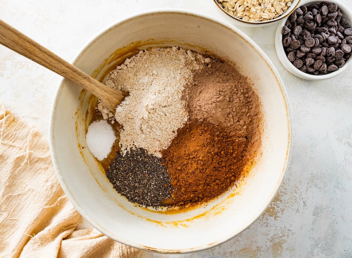 oat flour, cocoa powder, chocolate protein powder, chia seeds, baking soda, and salt being added to mixing bowl with wooden spoon to make healthy chocolate cookies.